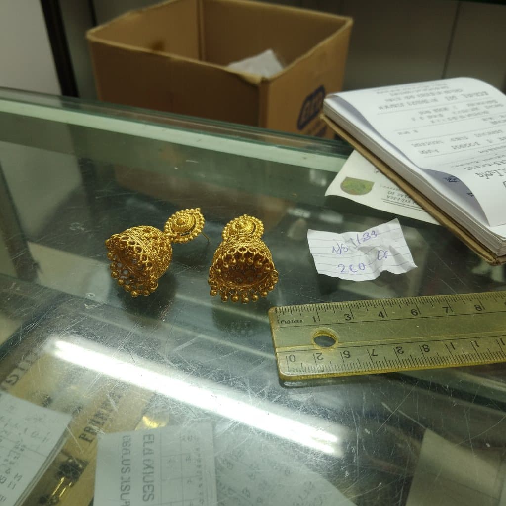 A hurried phone snap of gold earrings on a cluttered glass shop counter, next to a ruler and a paper price tag